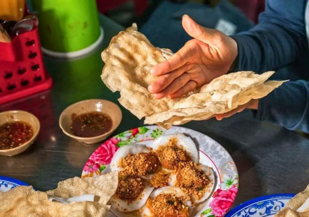 Đôi bàn tay đang bẻ miếng bánh tráng nướng giòn trên đĩa bánh đập phết mỡ hành và ruốc tôm bên chén mắm nêm